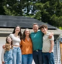 Happy Family Together In Front of Solar Equipped Home A Caucasian family with three children embrace on their driveway outside of their house. Their solar panel array is visible on the roof of their home, providing green natural energy for the environmentally conscious family. Sustainable modern lifestyle in action.